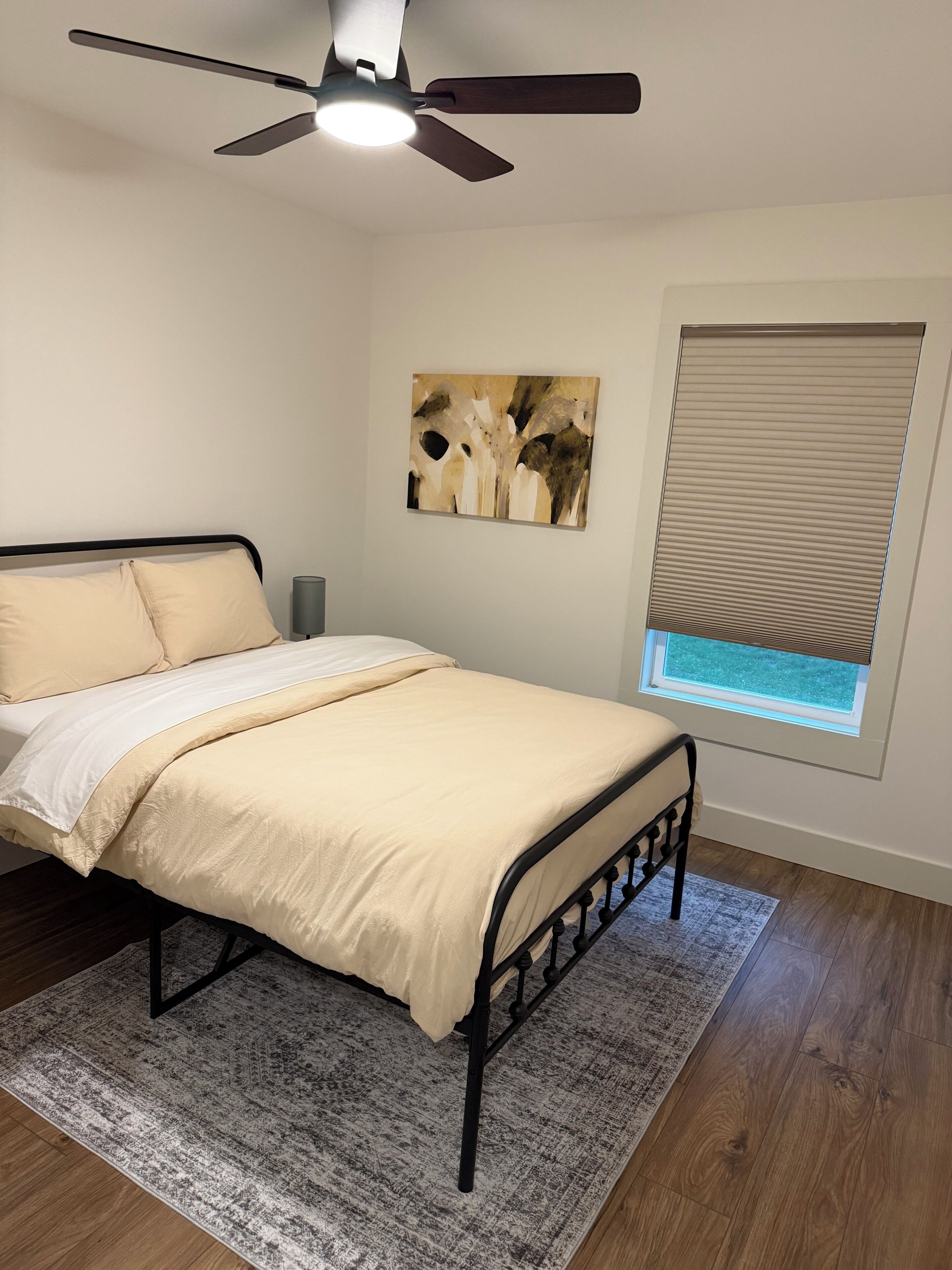 Um quarto com cama, ventilador de teto, janela com persianas e um quadro na parede.