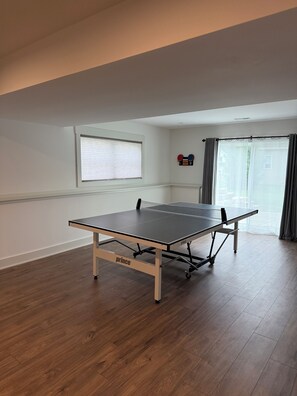 Een tafeltennistafel in een kamer met houten vloer, een raam met zonwering en een gordijn.