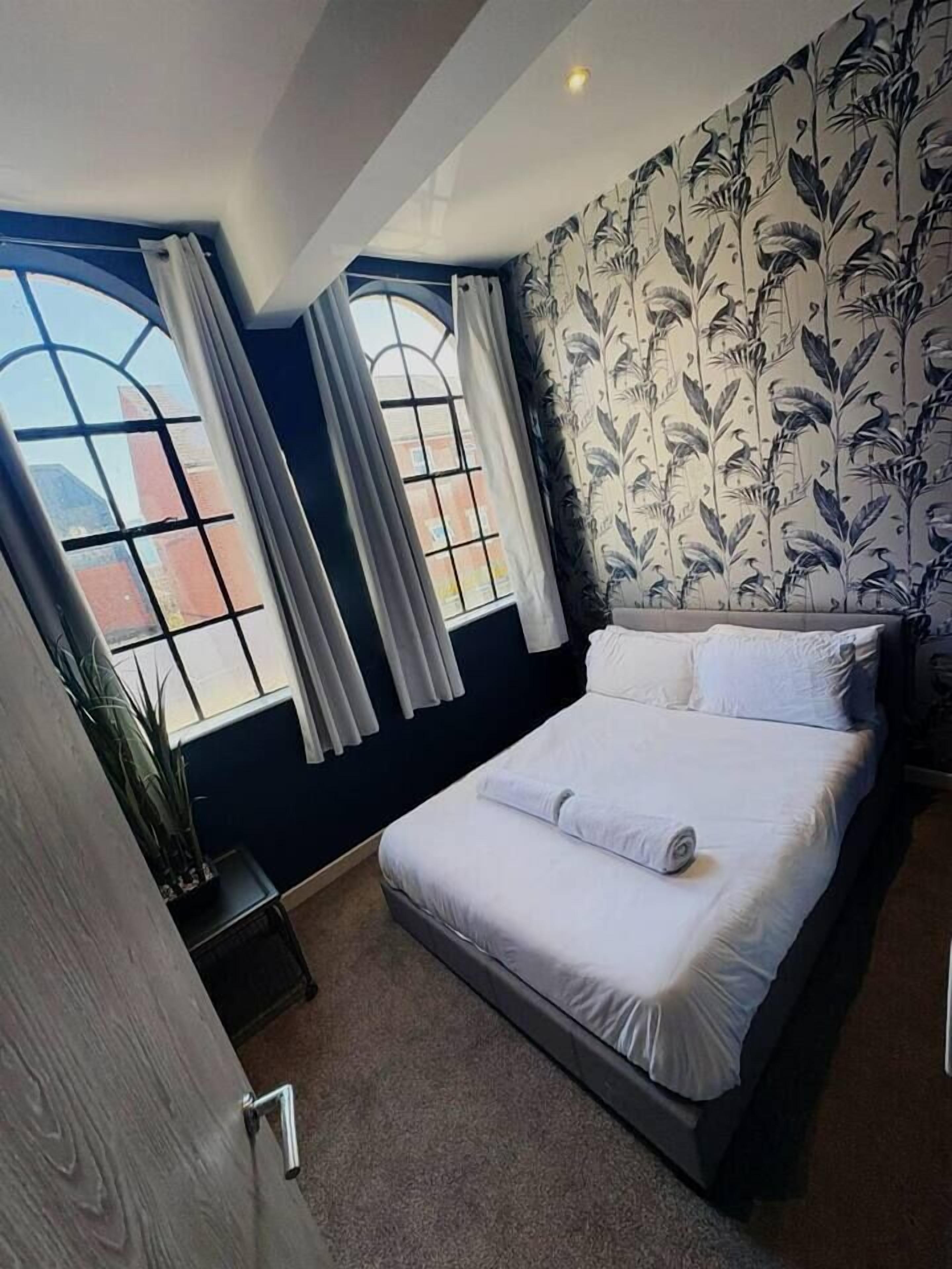 Hotelkamer met een groot bed, twee ramen met gordijnen, een kamerplant en een muur met bloemendesign.