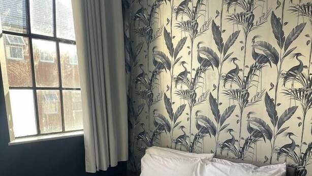 Ein ordentlich bezogenes Bett mit weißen Laken, ein Fenster mit Vorhängen und eine Wand mit floralem Wandpapier.