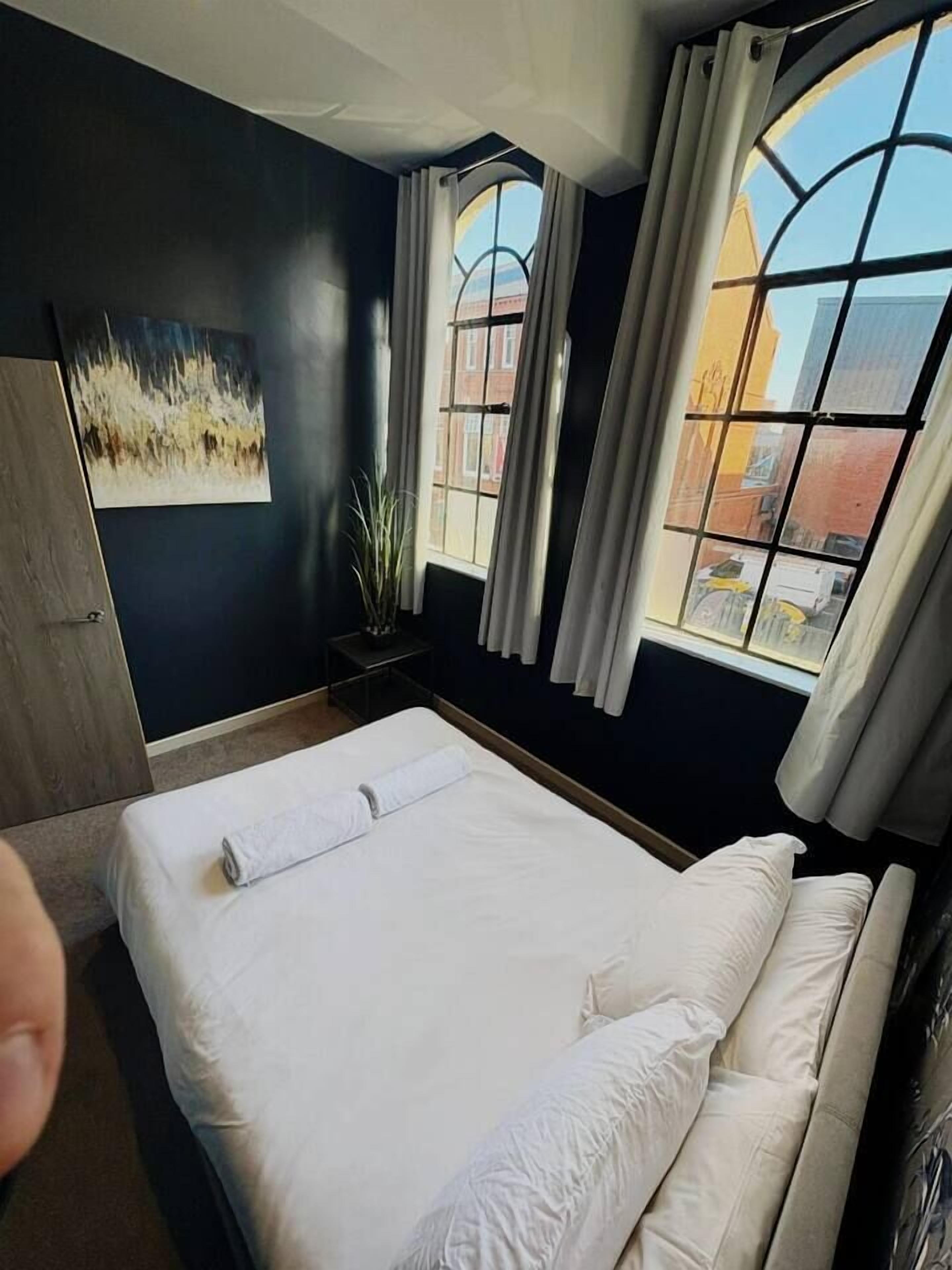 A hotel room with a large bed, a painting on the wall, a window with curtains, and a view of buildings outside.