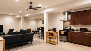Una sala de estar moderna con un sofá negro, un comedor con una mesa y sillas de madera, una cocina con gabinetes de madera oscura y electrodomésticos de acero inoxidable, y un ventilador de techo.
