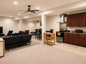 A modern living room with a black sofa, a dining area with a wooden table and chairs, a kitchen with dark wooden cabinets and stainless steel appliances, and a ceiling fan. - Spacious studio by the woods (Laurel)