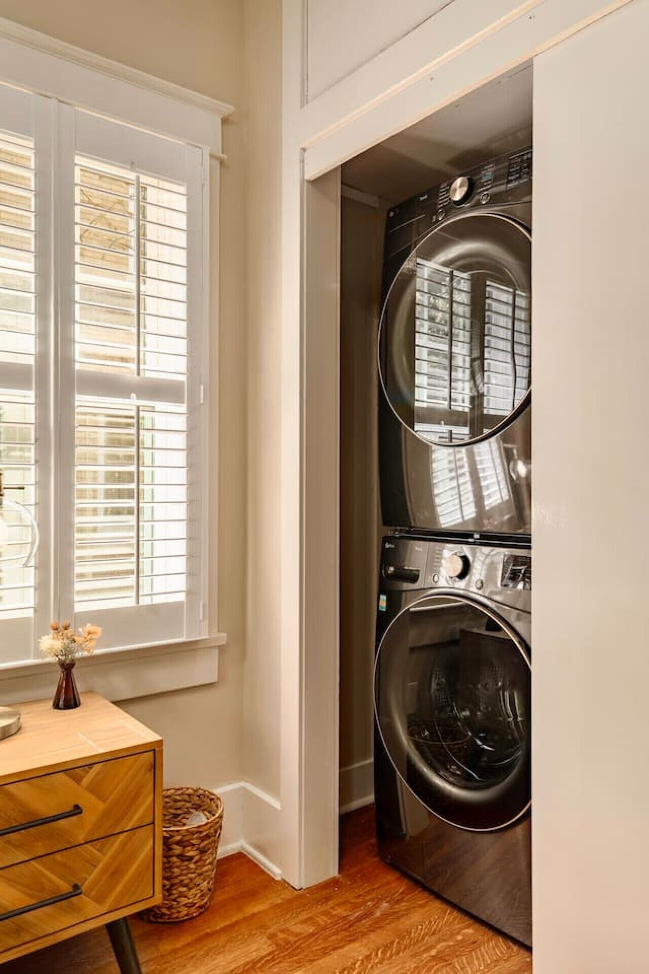 A laundry room with a double washer and dryer, a wooden cabinet, and a window with blinds.
