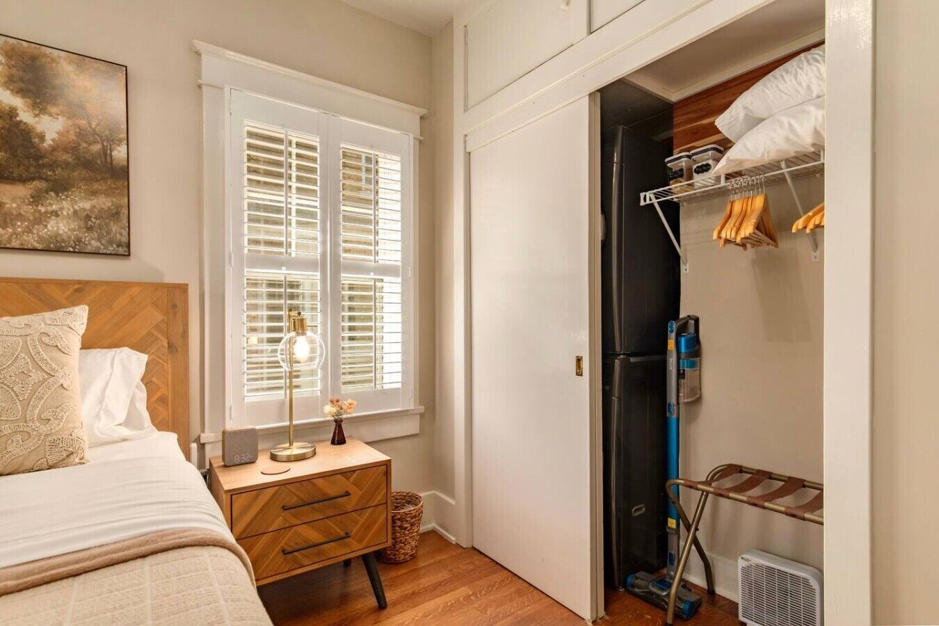 Une chambre à coucher avec un lit, une table de chevet, une lampe, une fenêtre avec des stores et un placard contenant des vêtements suspendus et un aspirateur.