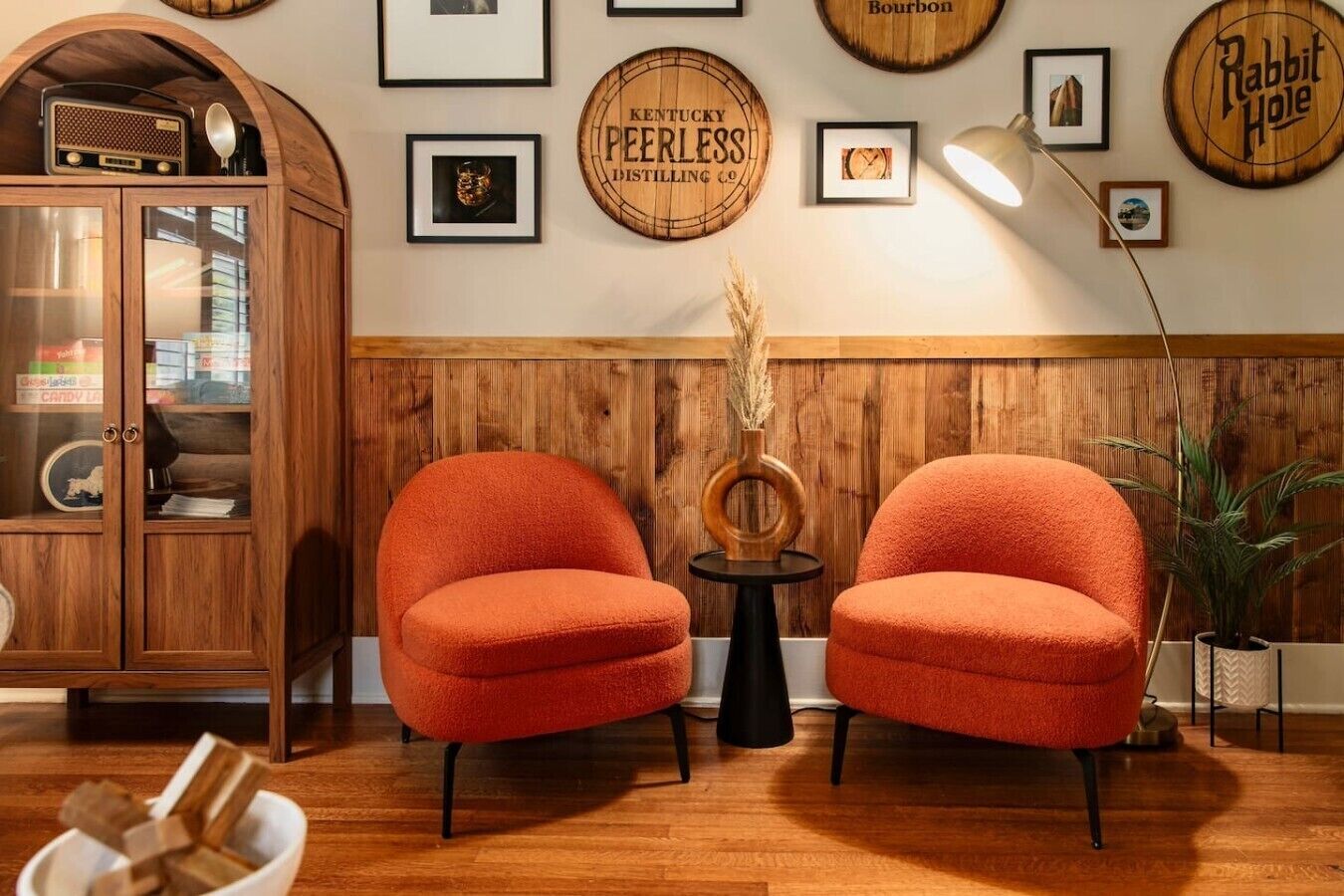 Een kamer met twee oranje fauteuils, een houten kast met glazen deuren, en een muur met diverse ingelijste foto’s en borden.