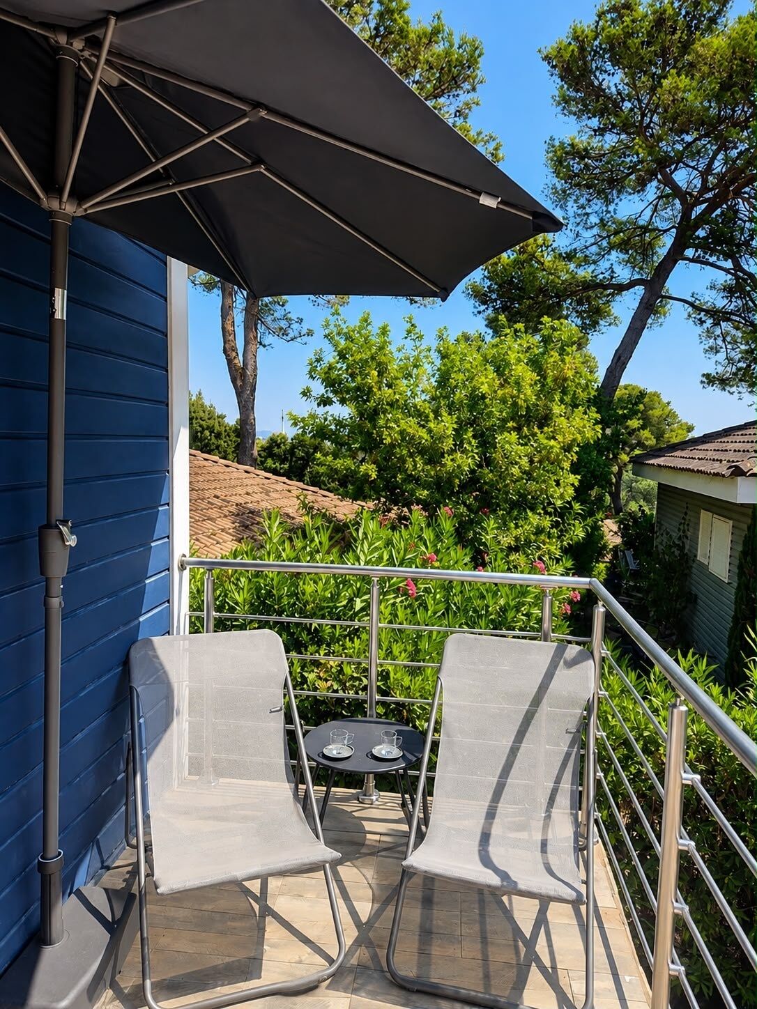 Een balkon met twee stoelen en een tafel, een zonnescherm en uitzicht op een tuin met bomen en een huis.