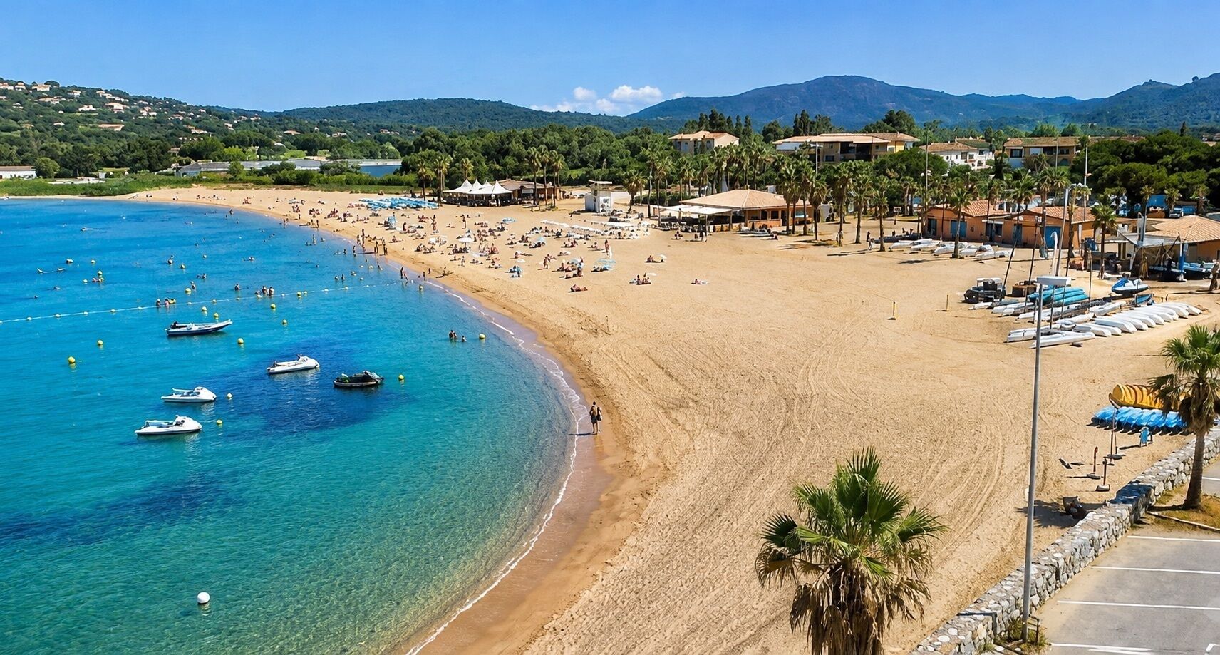 A beach with clear blue water, boats, and a sandy shore with umbrellas and buildings in the background.