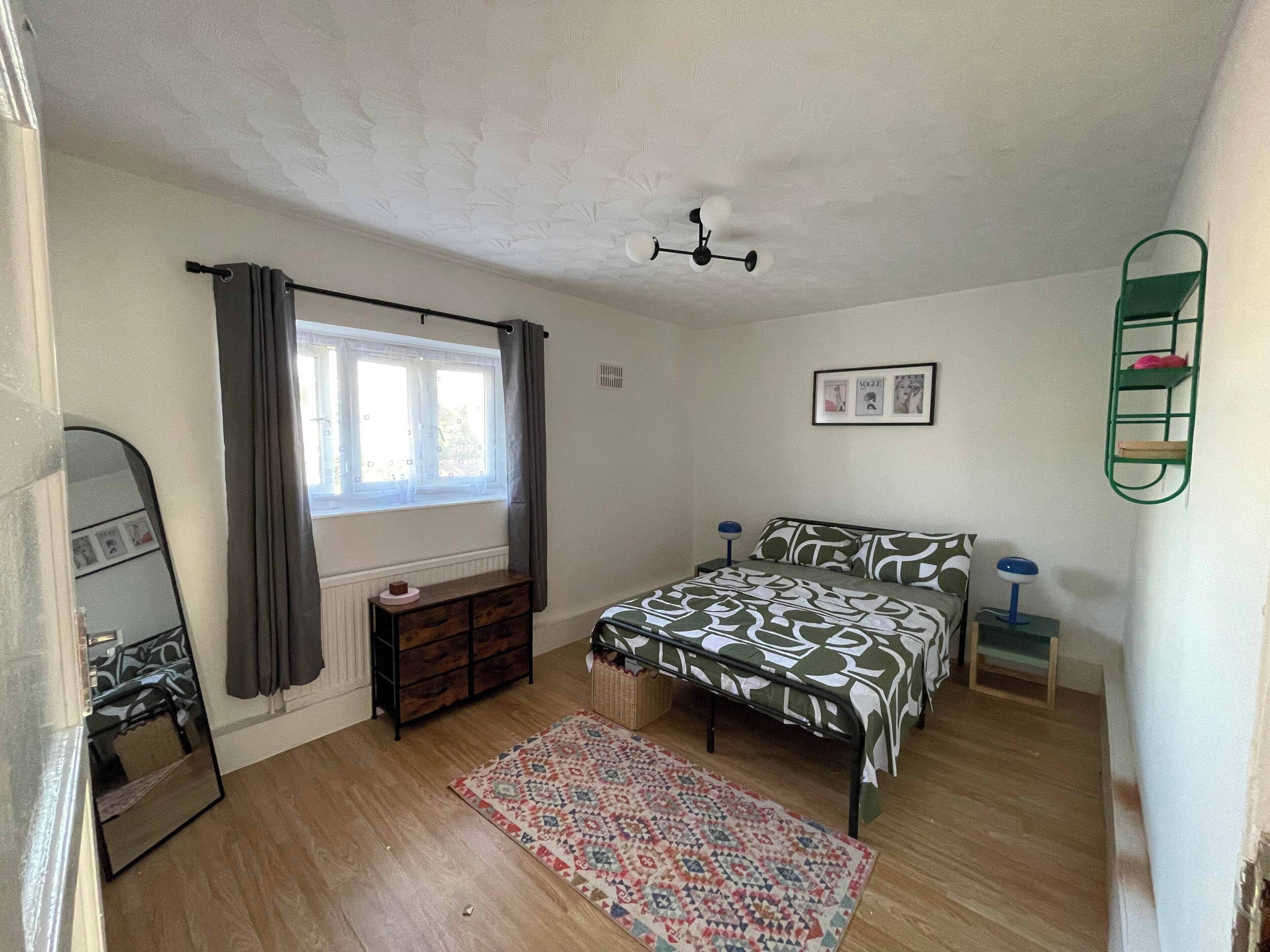 A bedroom with a bed, a dresser, a window with curtains, a ceiling fan, and a wall-mounted shelf.