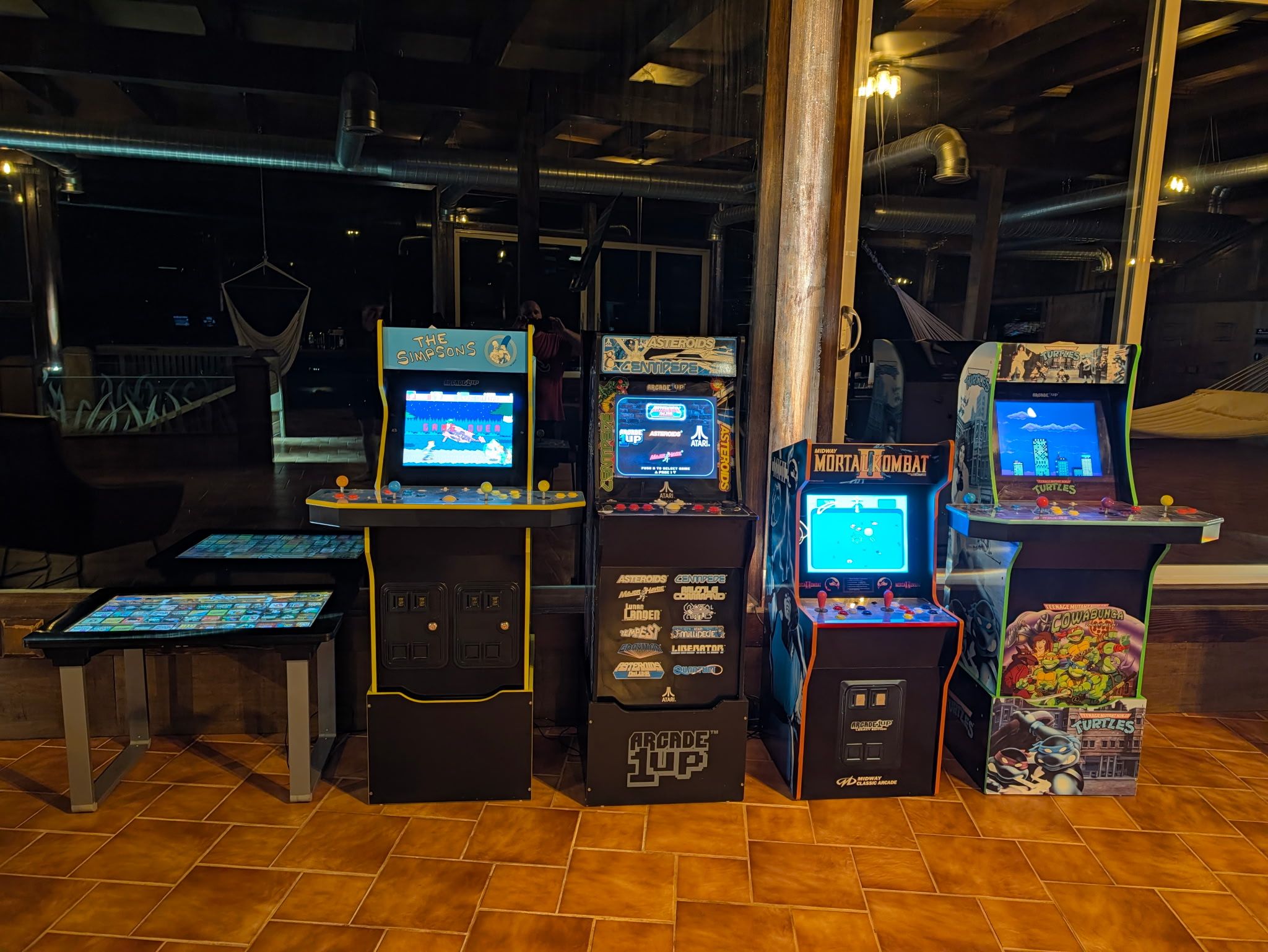 Uma fileira de máquinas de fliperama com diversos jogos, incluindo 'The Simpsons' e 'Mortal Kombat', em um ambiente interno com pouca luz.