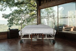 A bedroom with a large bed, wooden floor, a nightstand, and a window with a view of trees.