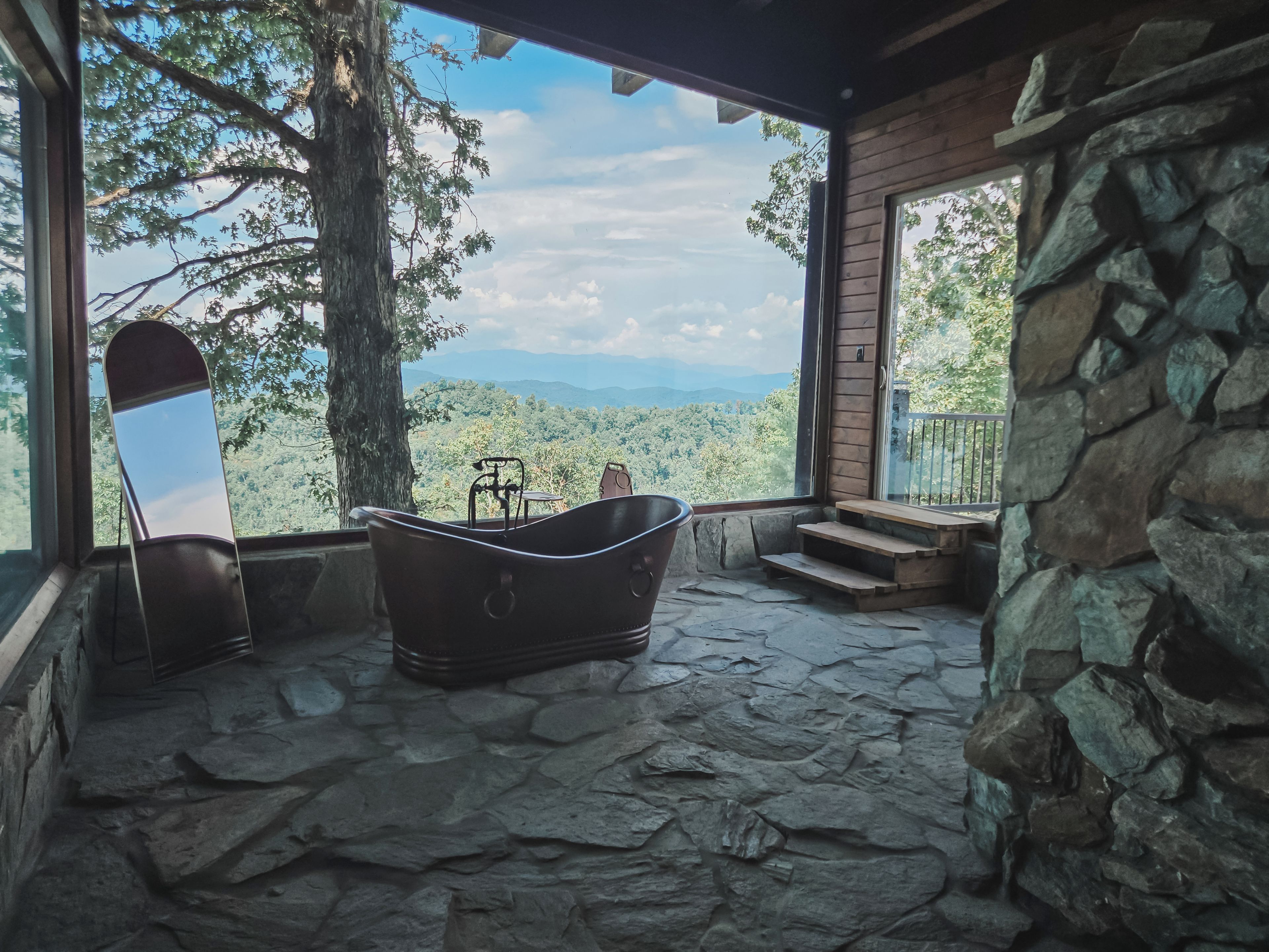 A stone-floored room with a bathtub, a chair, and a large window overlooking a forested landscape.