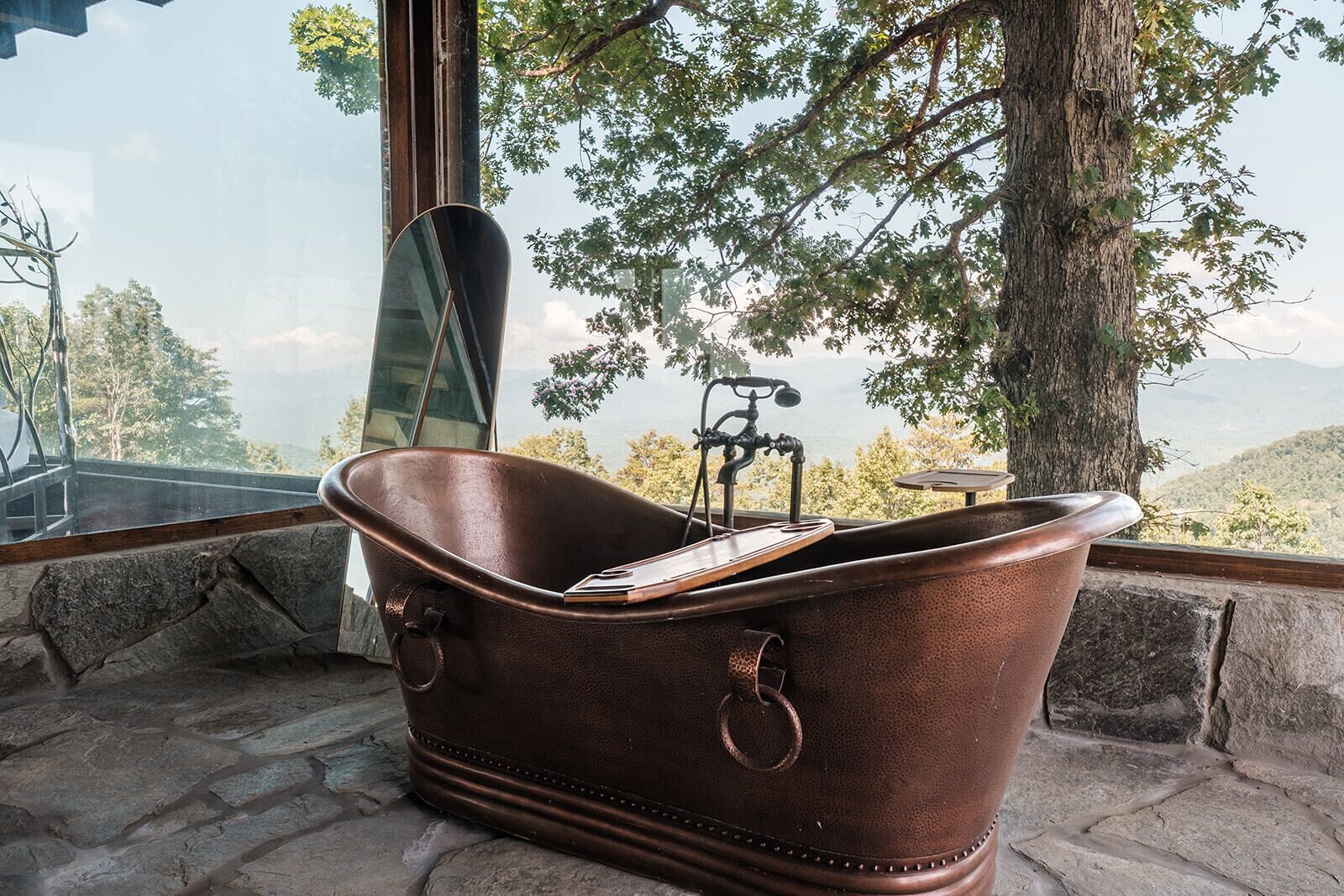 A bathtub with a smartphone inside, positioned by a window with a view of trees and mountains.