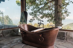 Una bañera con un teléfono inteligente en su interior, ubicada junto a una ventana con vistas a árboles y montañas.