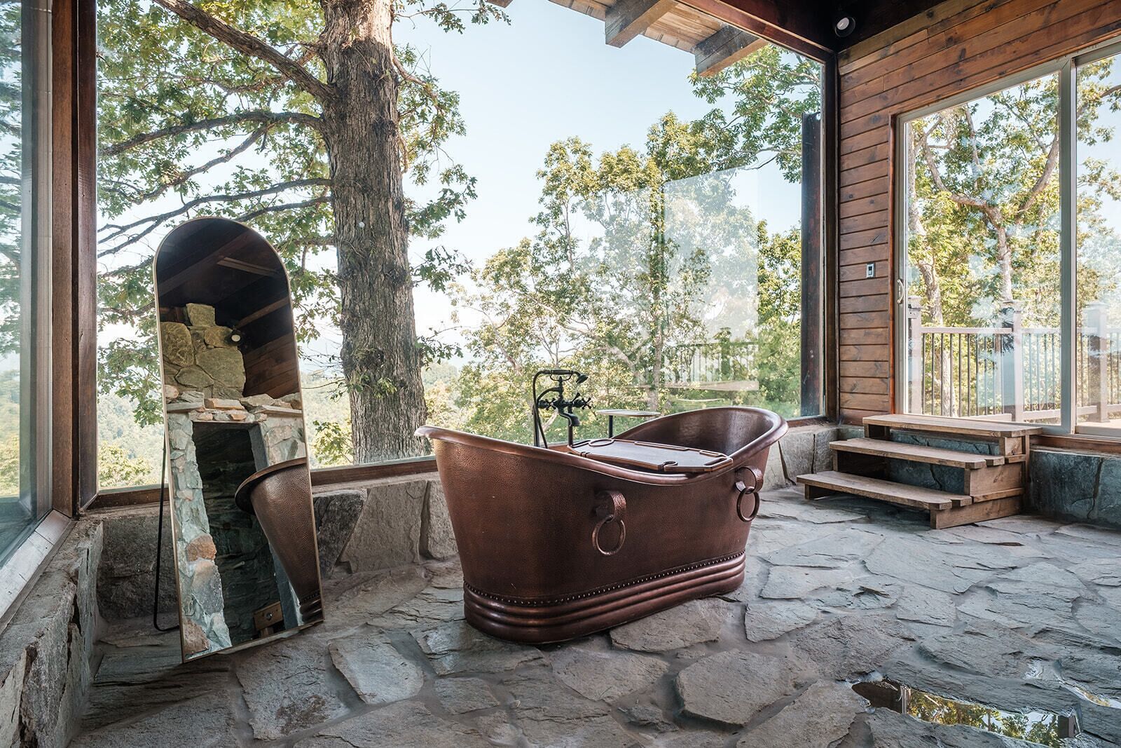 A stone-floored room with a large window, a stone fireplace, and a vintage bathtub.
