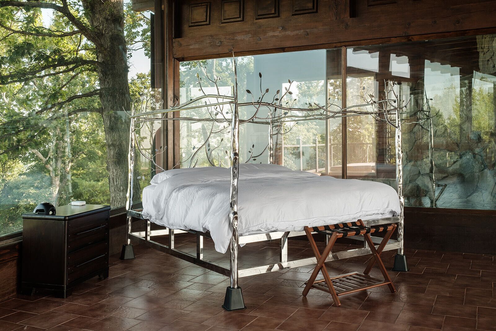 A bed with a metal frame and white bedding, a wooden nightstand, and a view of trees through large windows.