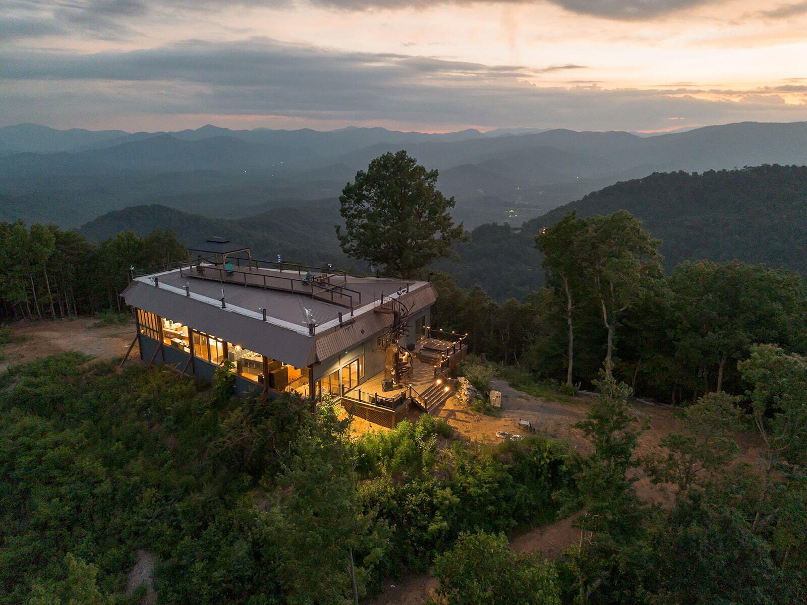 Uma casa moderna com telhado plano, grandes janelas e um deck, rodeada por uma floresta densa e montanhas.
