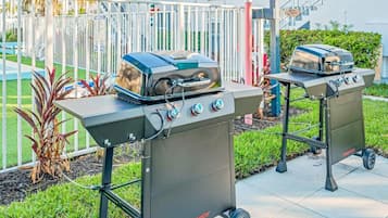 Deux barbecues portables de la marque Megamaster, équipés de roulettes, installés à l’extérieur, avec une clôture blanche et de la verdure en arrière-plan.