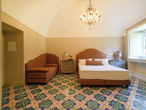 Un dormitorio con una cama grande, un sillón marrón, una mesita de noche con lámpara y una araña.