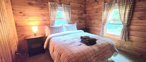 Une chambre dans un chalet en bois, avec un lit, une table de chevet et deux fenêtres avec des rideaux.
