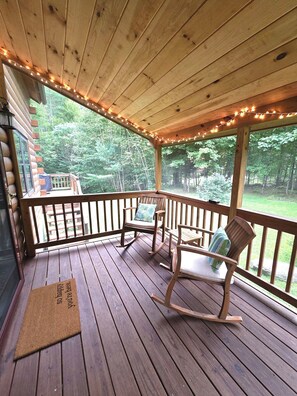 A wooden deck with a rocking chair and a bench, a doormat, and a view of the surrounding forest.
