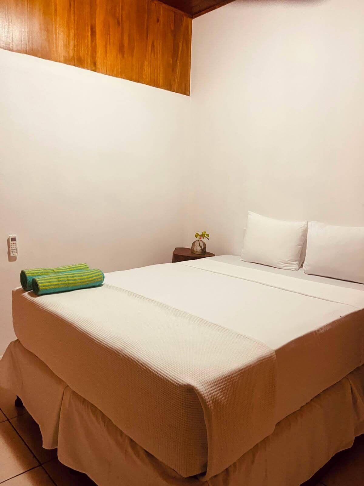 Habitación individual con techo de madera, una mesita de noche con un florero y una toalla verde sobre la cama.