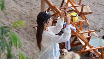 Una persona de pie en una playa arenosa con un perro, una estructura de sillas de madera y un bolso amarillo.