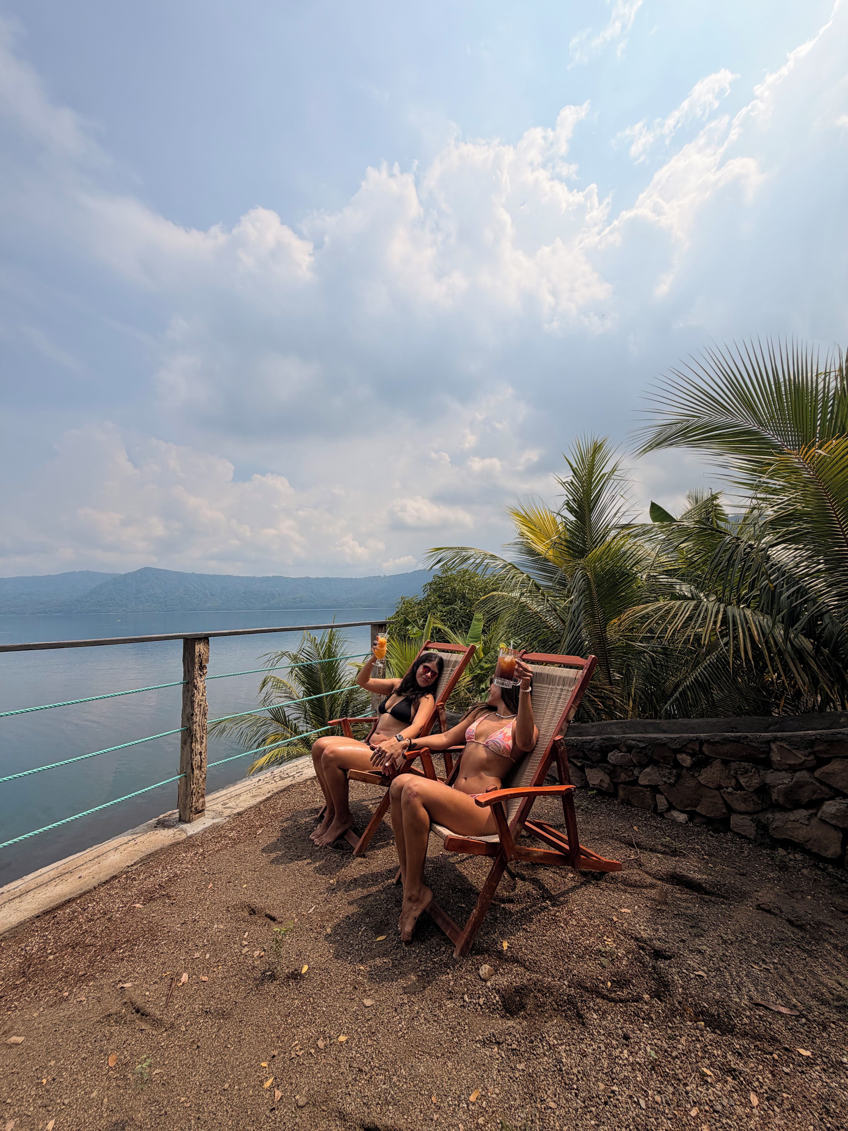 Dos personas sentadas en sillas de madera junto a una piscina, con vista al mar y a las montañas de fondo.