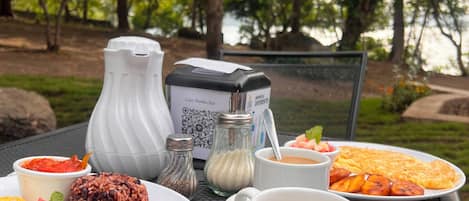 En opsætning med mad på et udendørs bord, inklusive en QR-kode, en kande vand, krydderier og en kop kaffe.