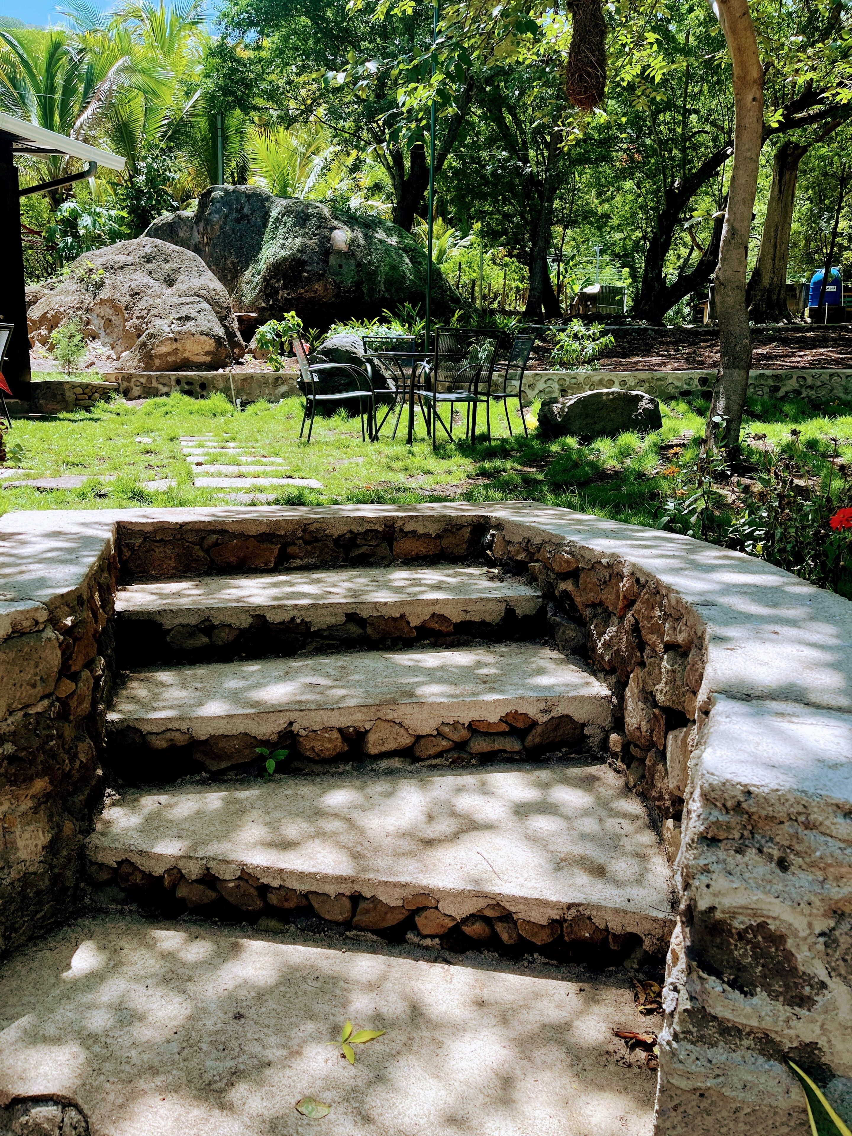 A set of stone steps leading to a seating area with metal chairs, surrounded by greenery and large rocks.