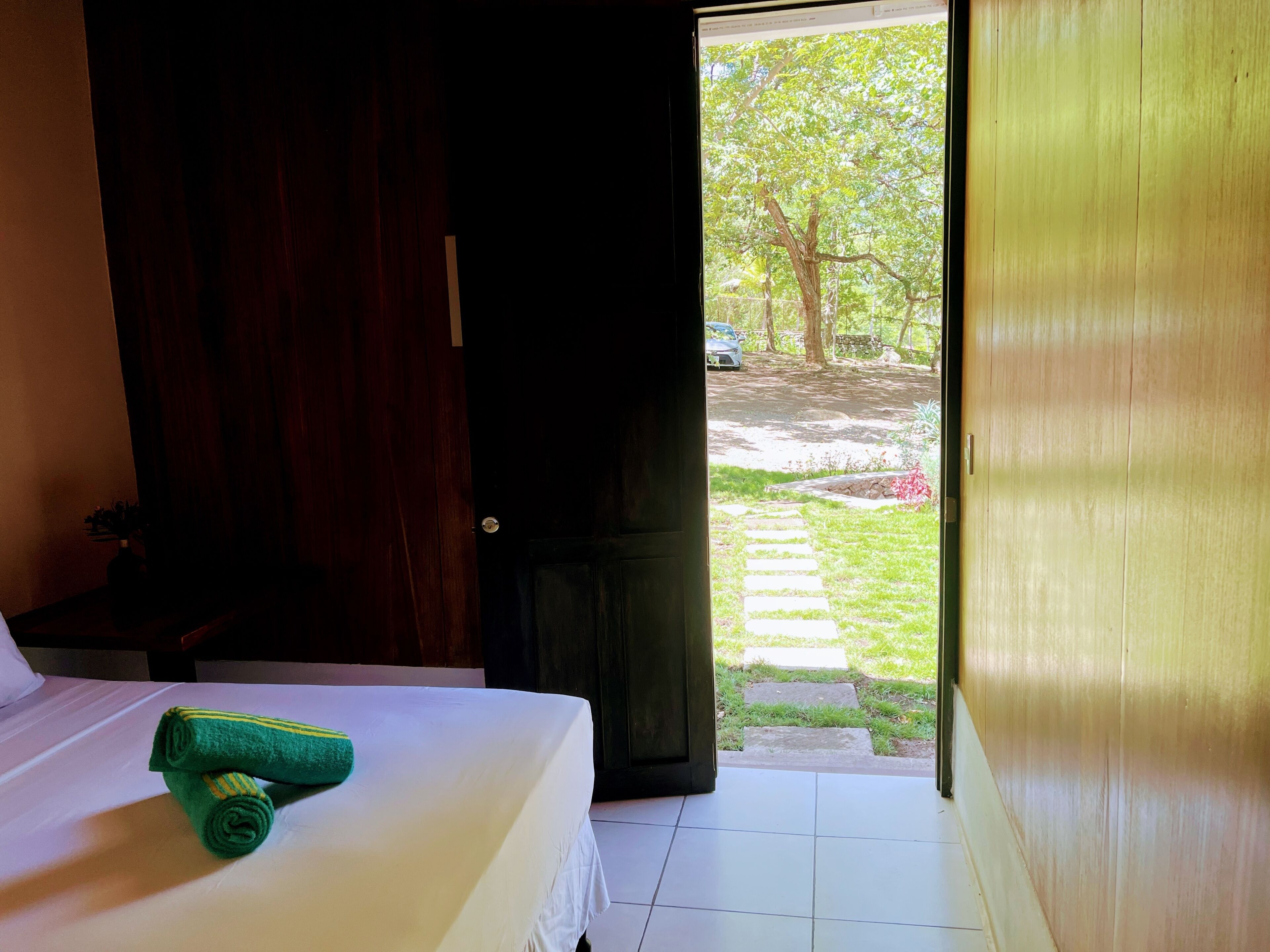 Quarto de hotel com cama, porta de madeira e vista para um jardim com caminho.