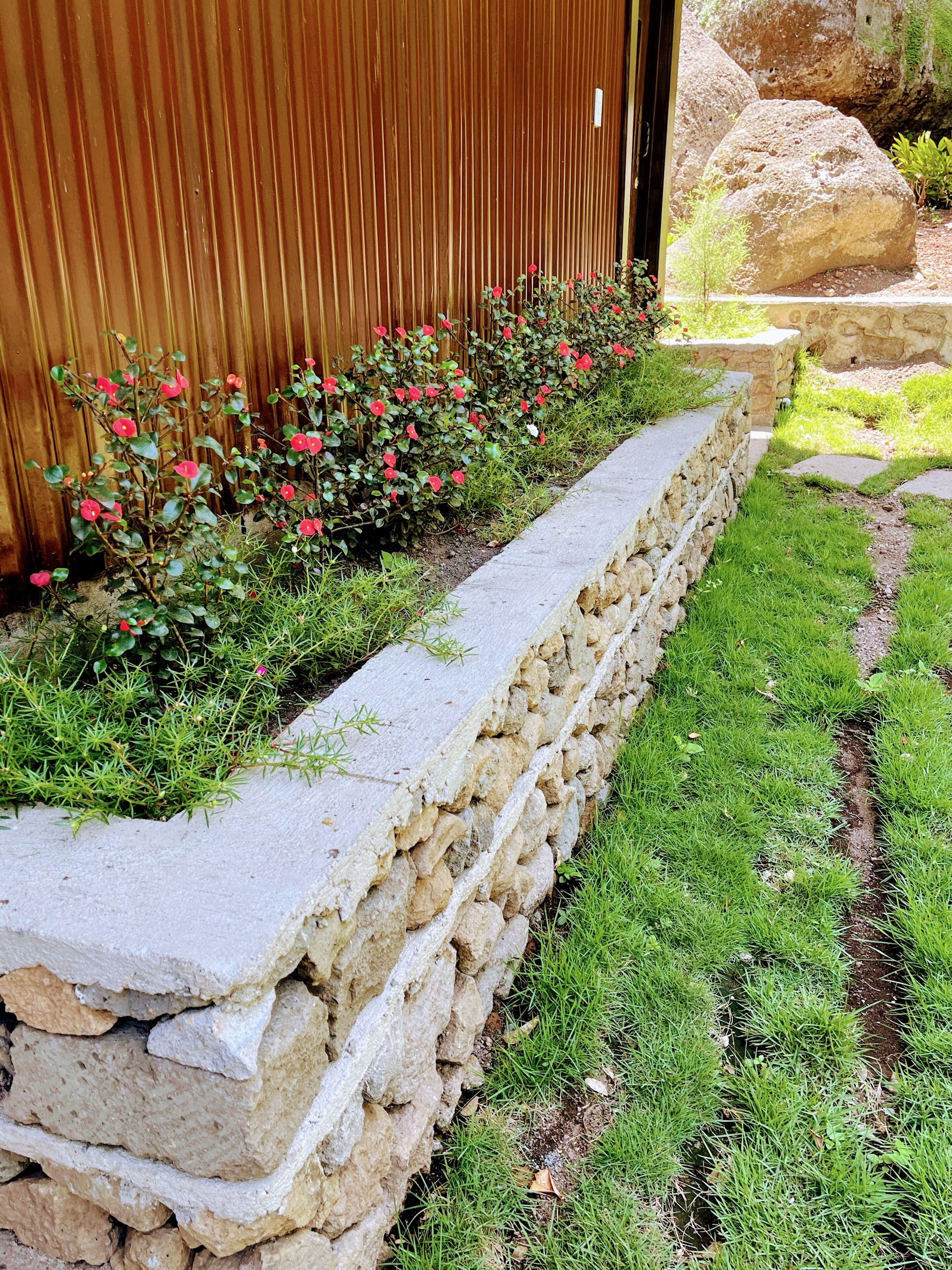A stone retaining wall with a grassy garden area and a wooden fence in the background.