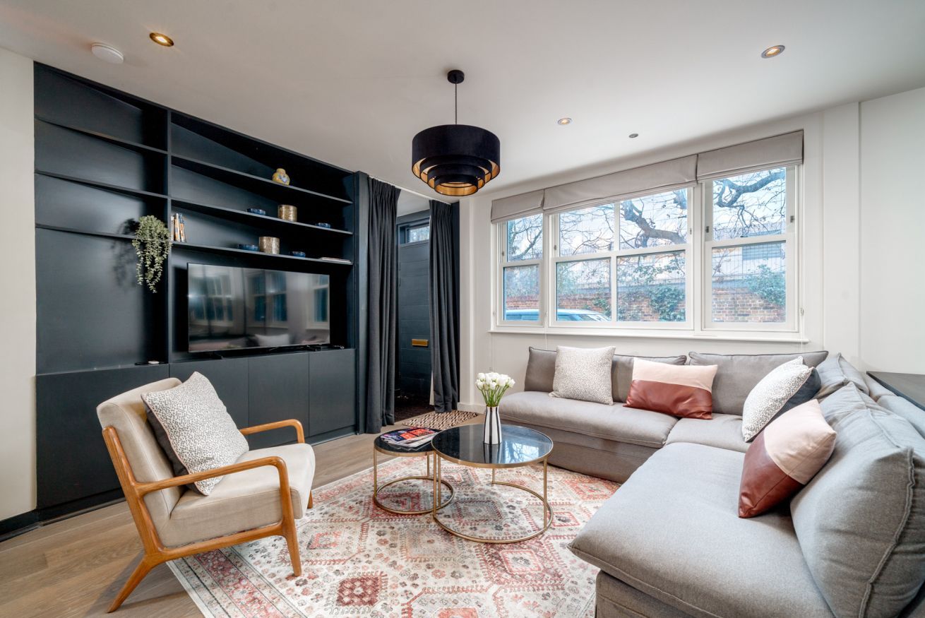 Uma sala de estar moderna com sofá, poltrona e mesa de centro. Há uma janela grande com vista para árvores e um muro de tijolos.