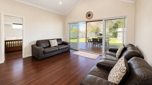 A spacious living room with a dark brown leather sofa, a wooden bench, and a sliding glass door leading to an outdoor patio. - Driftwood Cottage – Coastal Comfort in Central Busselton (Busselton)