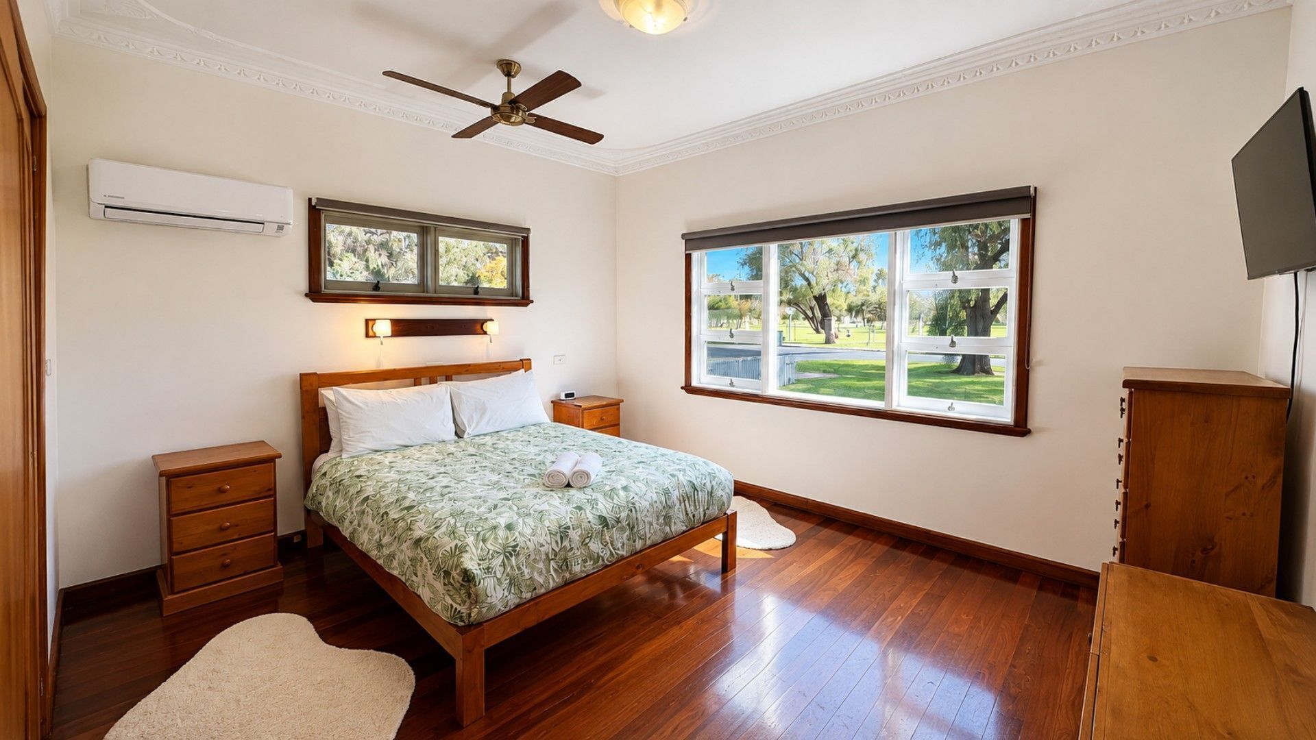 A bedroom with a bed, a nightstand, a wooden dresser, a ceiling fan, and a window with a view of a park.