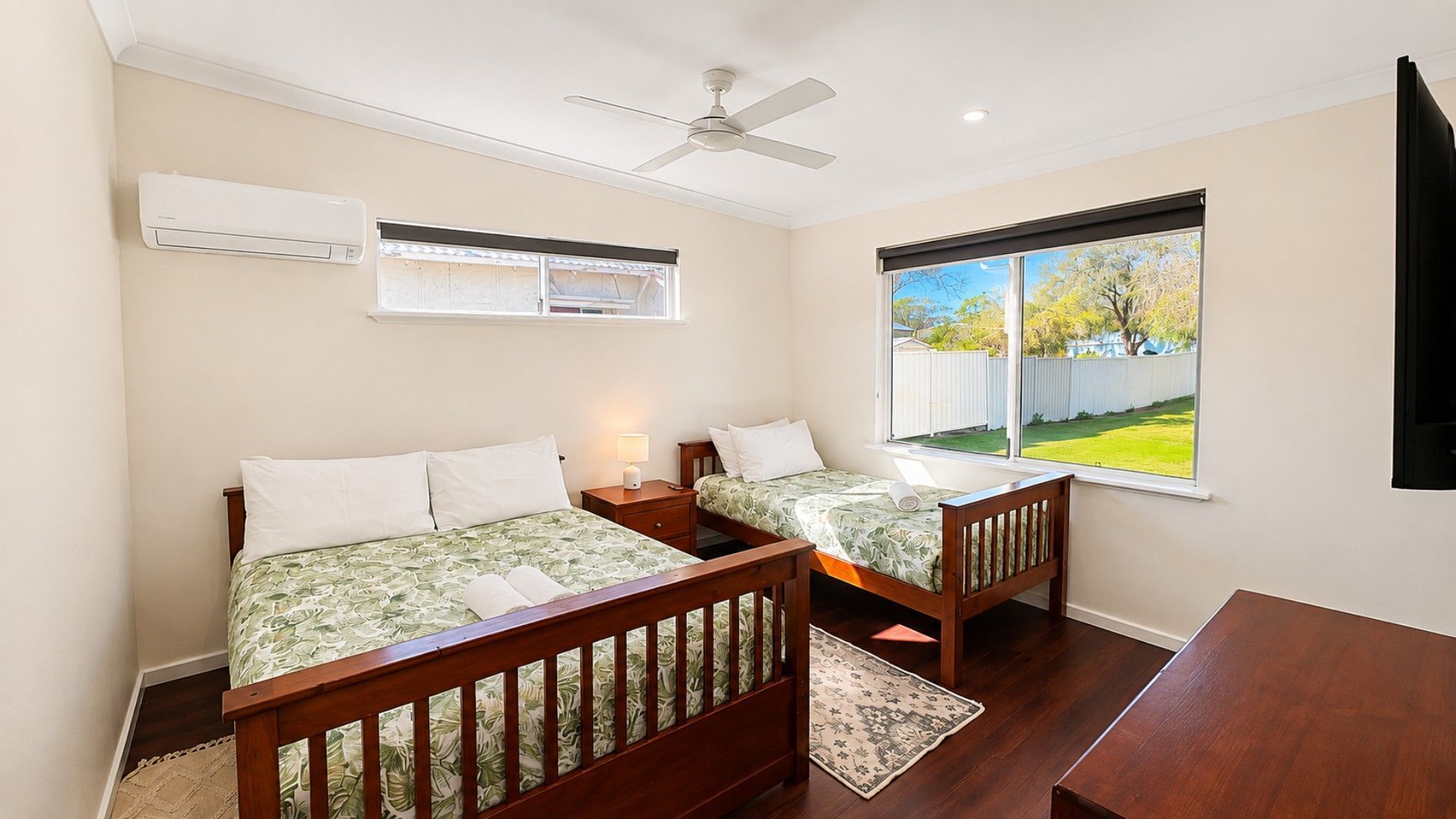 A bedroom with two beds, a ceiling fan, an air conditioning unit, and a window with a view of a backyard.