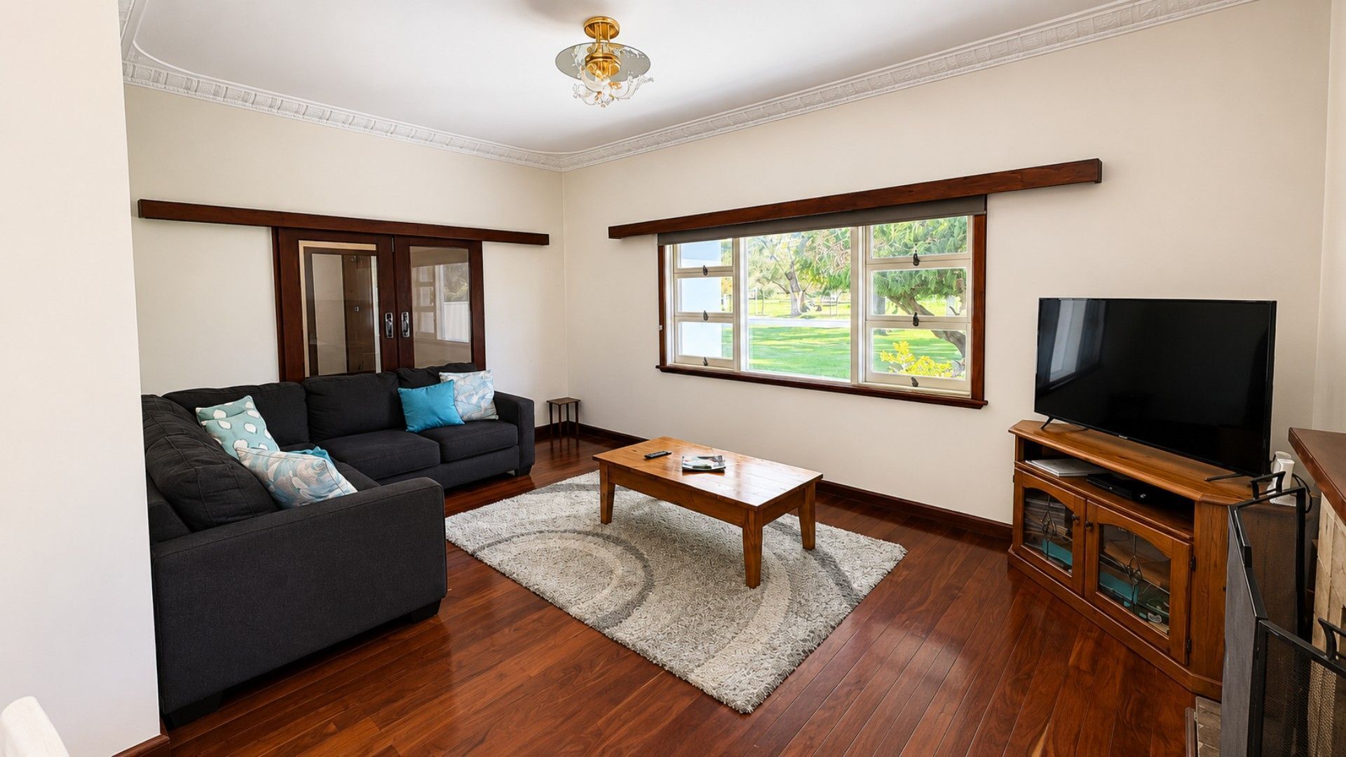A living room with a dark-colored sofa, a wooden coffee table, a television on a wooden stand, and a large window with a view of greenery.