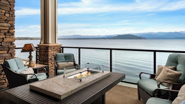 An outdoor patio with a stone fireplace, seating, and a view of a lake and mountains. - Waterfront Townhouse, Private Hot Tub, Boat Slip (Sandpoint)