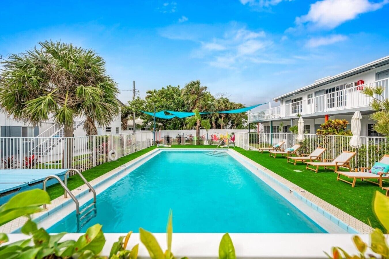 A swimming pool with a white fence, lounge chairs, and a building with balconies.