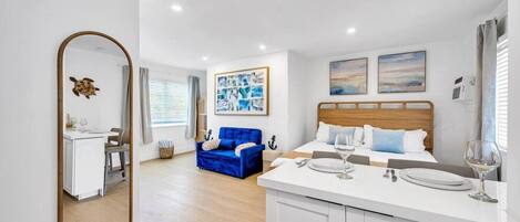 A modern hotel room with a bed, a seating area with a blue sofa, a dining table with a white tablecloth, and a large mirror.