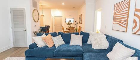 A living room with a blue sectional sofa, a coffee table, and a dining area in the background.