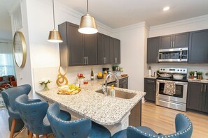 A modern kitchen with granite countertops, dark cabinets, and stainless steel appliances. There are blue chairs around the island and pendant lights above. - Lux Master bedroom in a 3 bed 2 bath house (Charlotte)