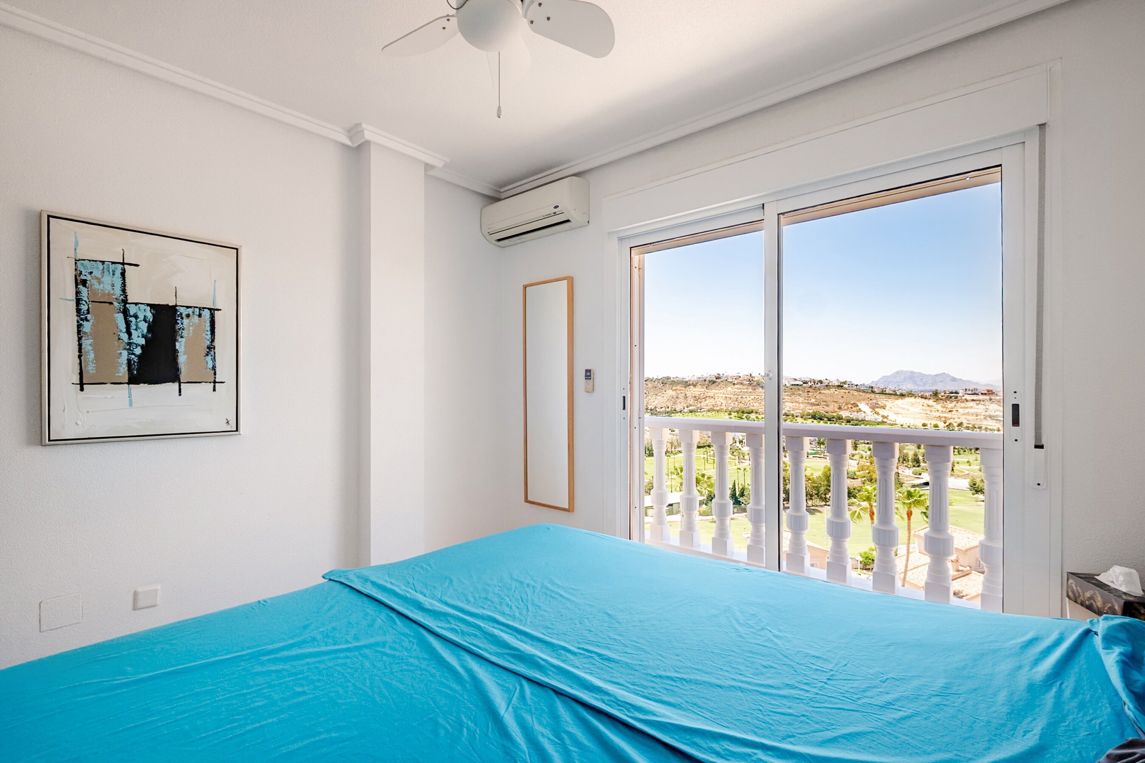 Une chambre avec une grande fenêtre, un ventilateur de plafond et un tableau accroché au mur.