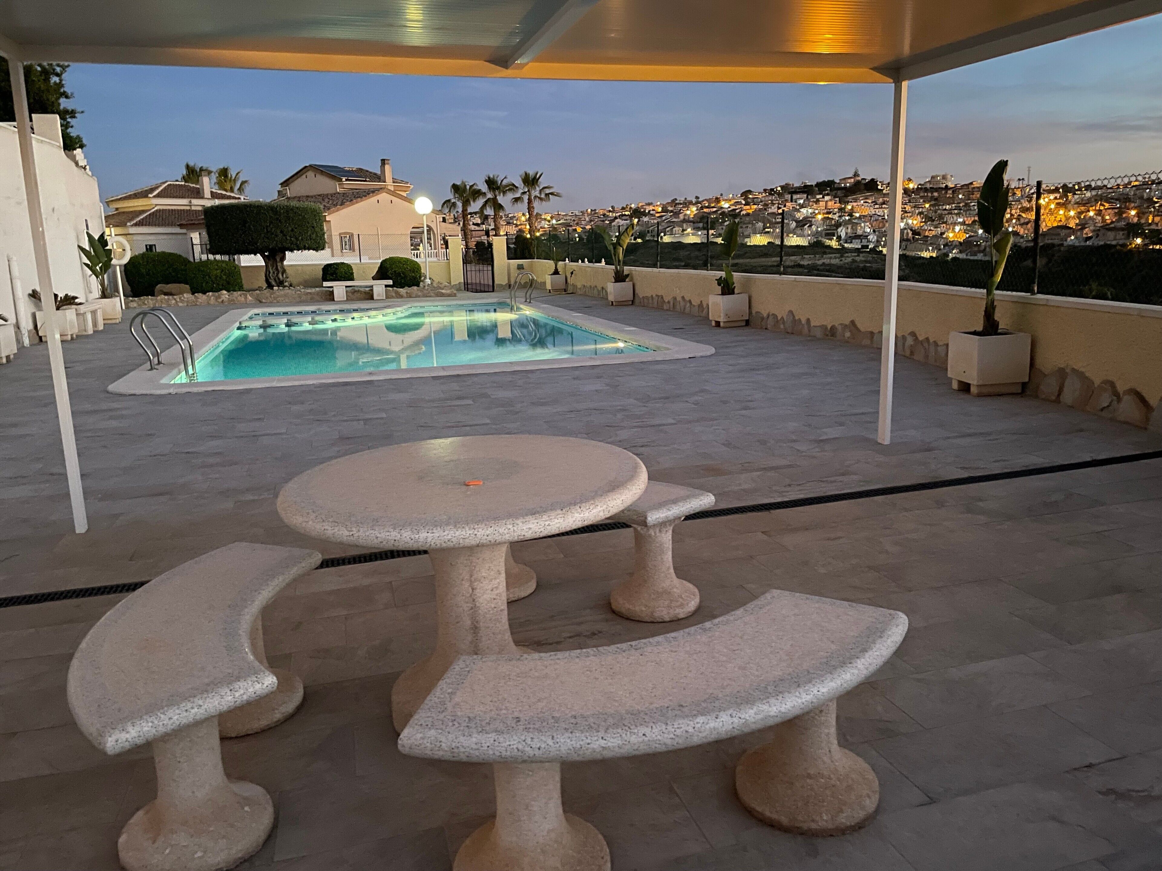 Un espace extérieur avec une piscine, une table et des bancs, entouré d’un petit muret et offrant une vue sur le paysage urbain au crépuscule.
