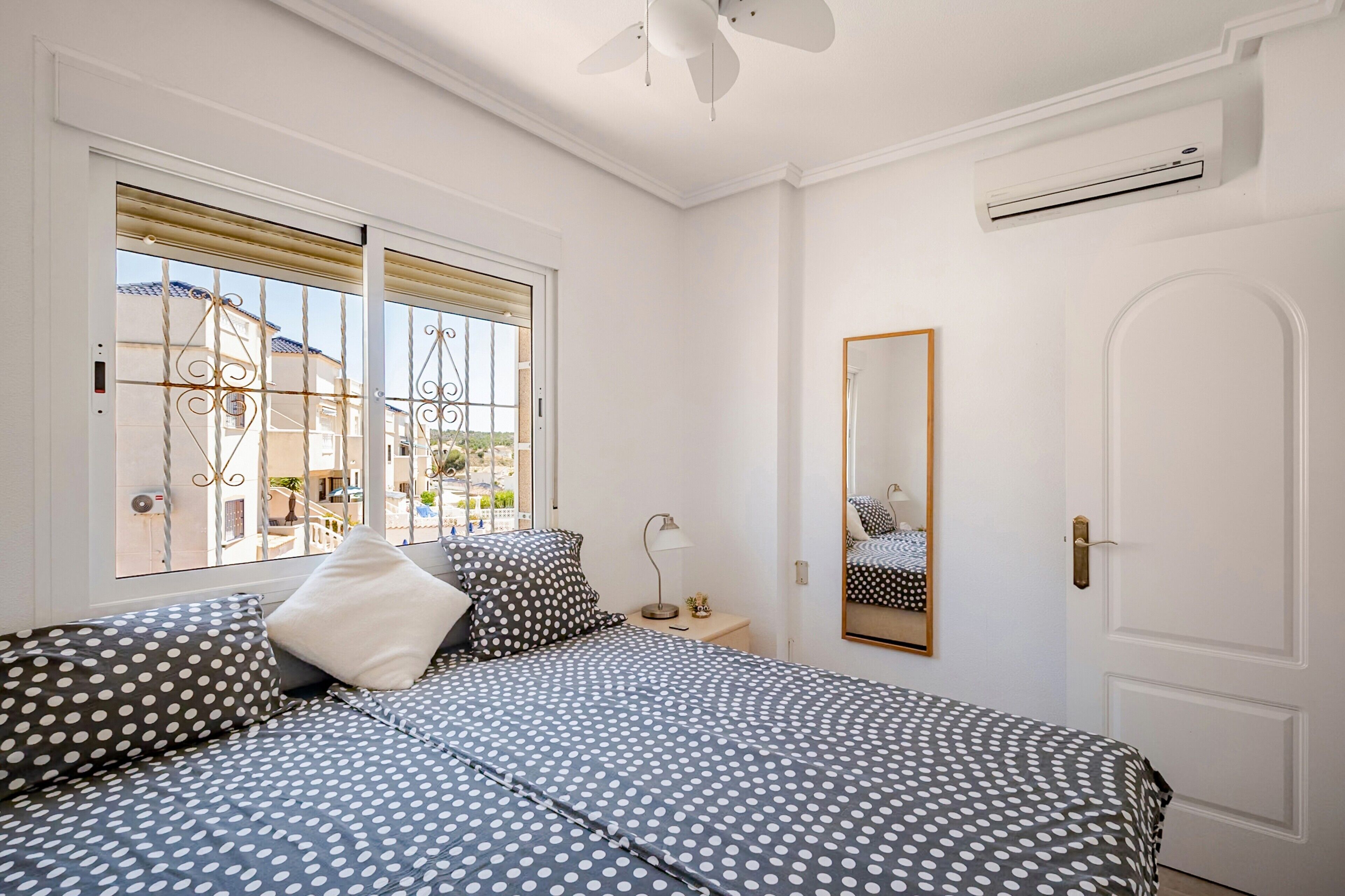 A bedroom with a bed, a bedside table, a lamp, a mirror, and a window with a view of buildings outside.