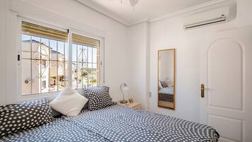 A bedroom with a bed, a bedside table, a lamp, a mirror, and a window with a view of buildings outside.