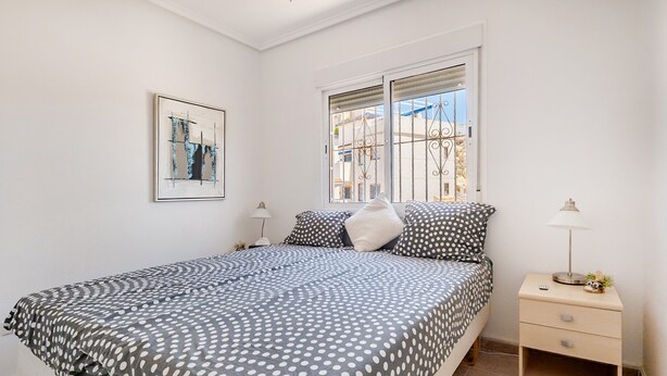 A bedroom with a bed, a nightstand, a lamp, a window with bars, and a framed picture on the wall.