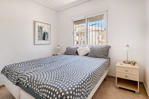 A bedroom with a bed, a nightstand, a lamp, a window with bars, and a framed picture on the wall.