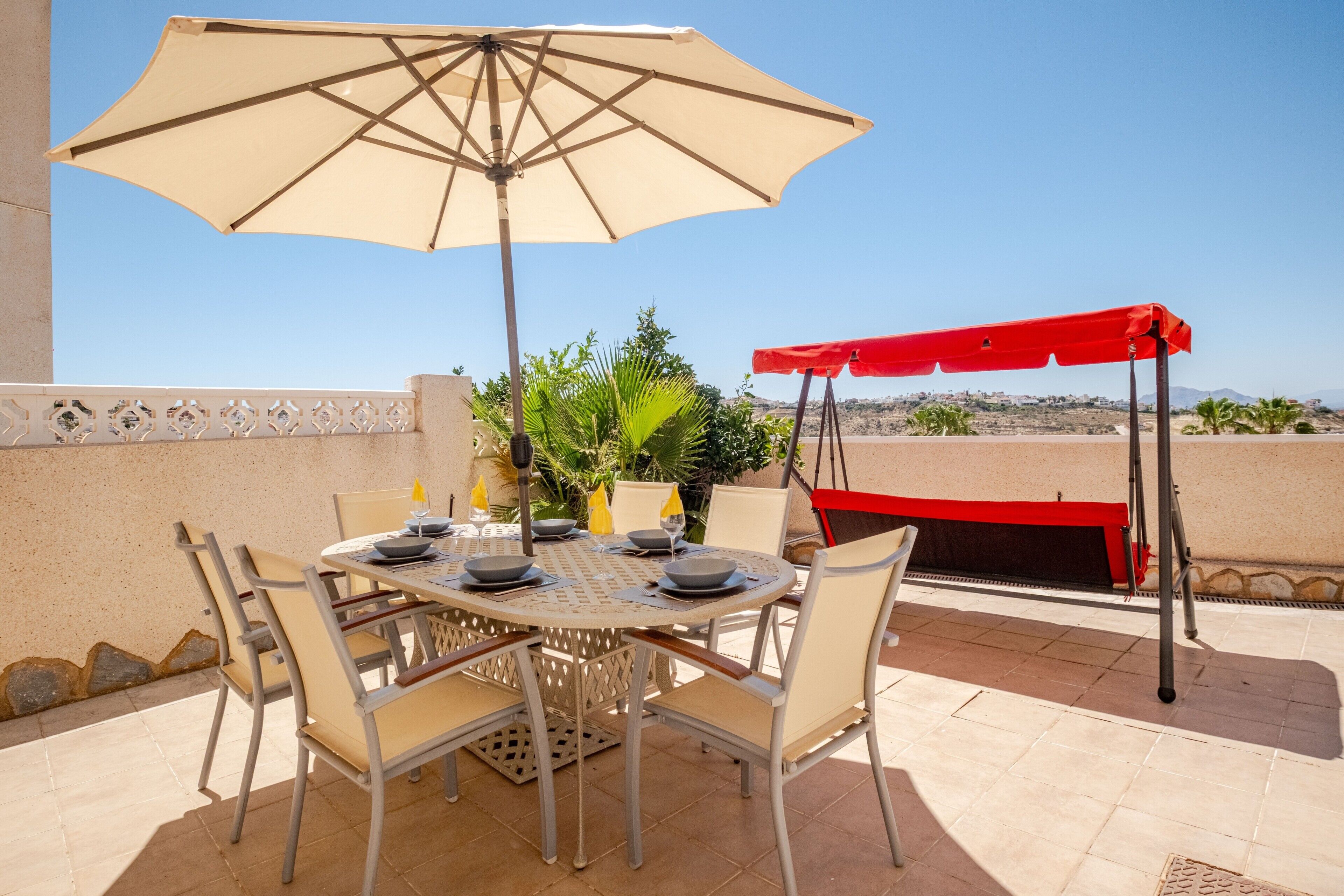 Une terrasse extérieure avec une table ronde dressée pour le repas, entourée de chaises, et un grand parasol offrant de l’ombre.