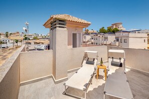 Eine Dachterrasse mit zwei Liegestühlen, einem kleinen Tisch, einer Flasche und einem Glas, und Blick auf die umliegenden Gebäude.